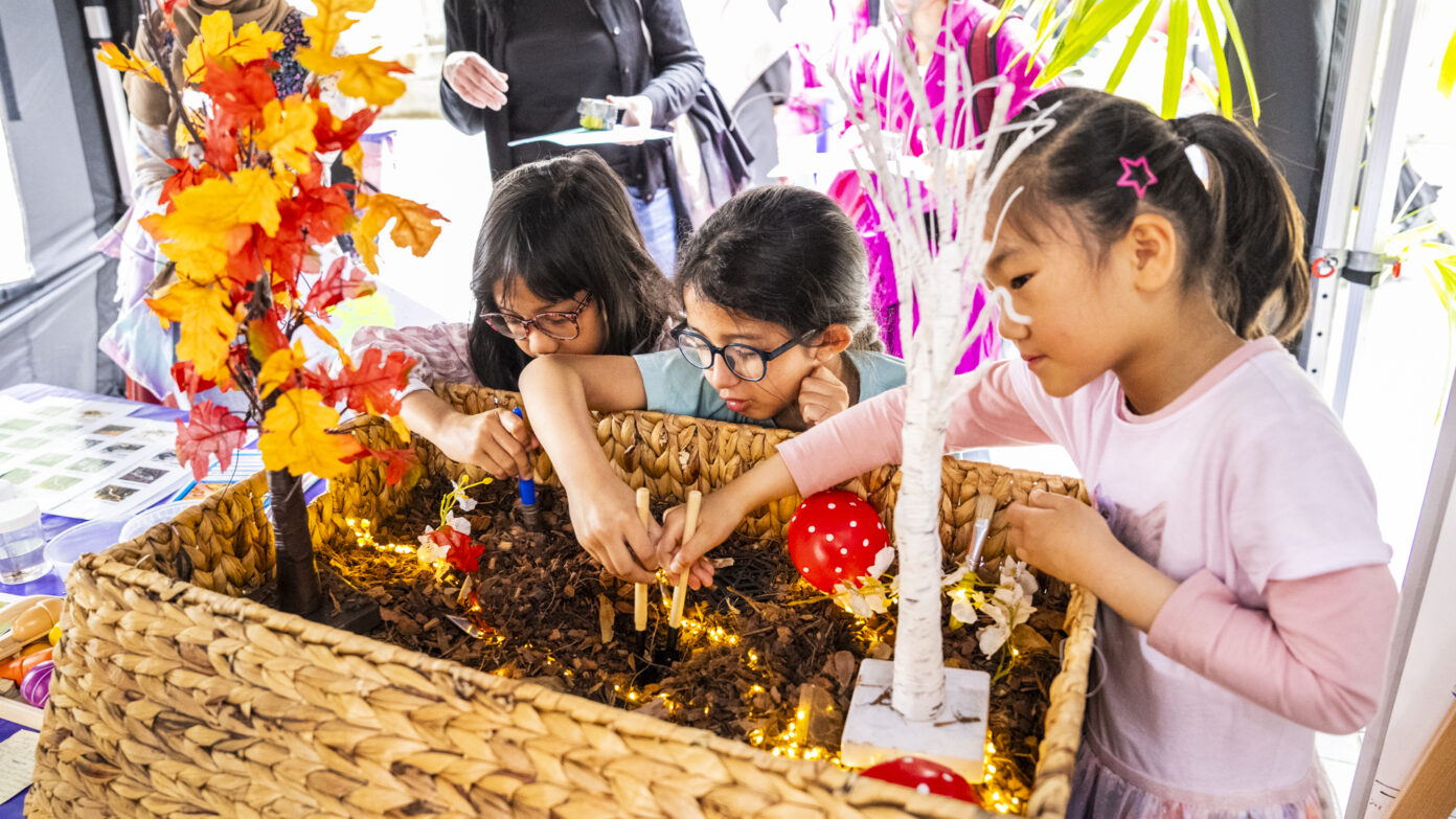 Three children exploring a sensory activity with soil, small trees, and lights in a woven basket