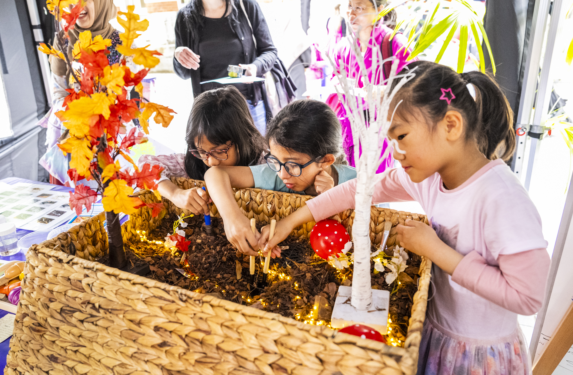 Three children exploring a sensory activity with soil, small trees, and lights in a woven basket