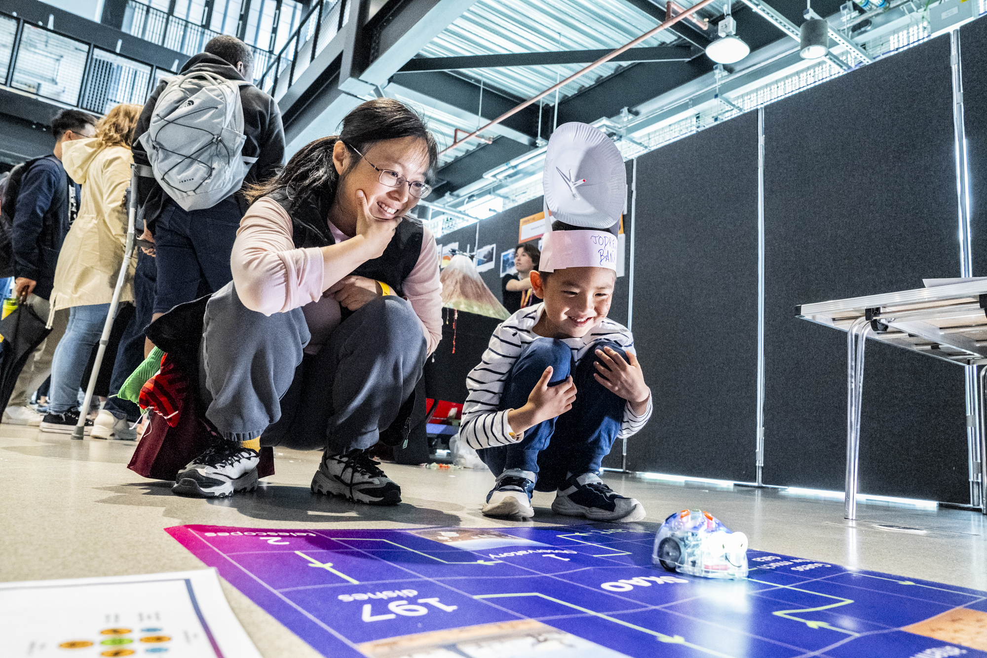 Young boy in striped top with telescope paper hat and women in peach top watch robot moving across a mat