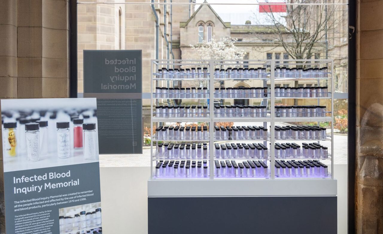 A glass display titled “Infected Blood Inquiry Memorial,” showing rows of small glass bottles arranged on illuminated shelves inside a building, with an explanatory panel to the left and a stone courtyard and historic buildings visible through the window behind the installation.